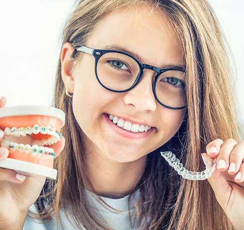 smiling girl holding up a set of clear aligners and a 3D model of a set of teeth with braces