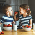 a brother and sister smile as they eat strawberries and milk in the kitchen
