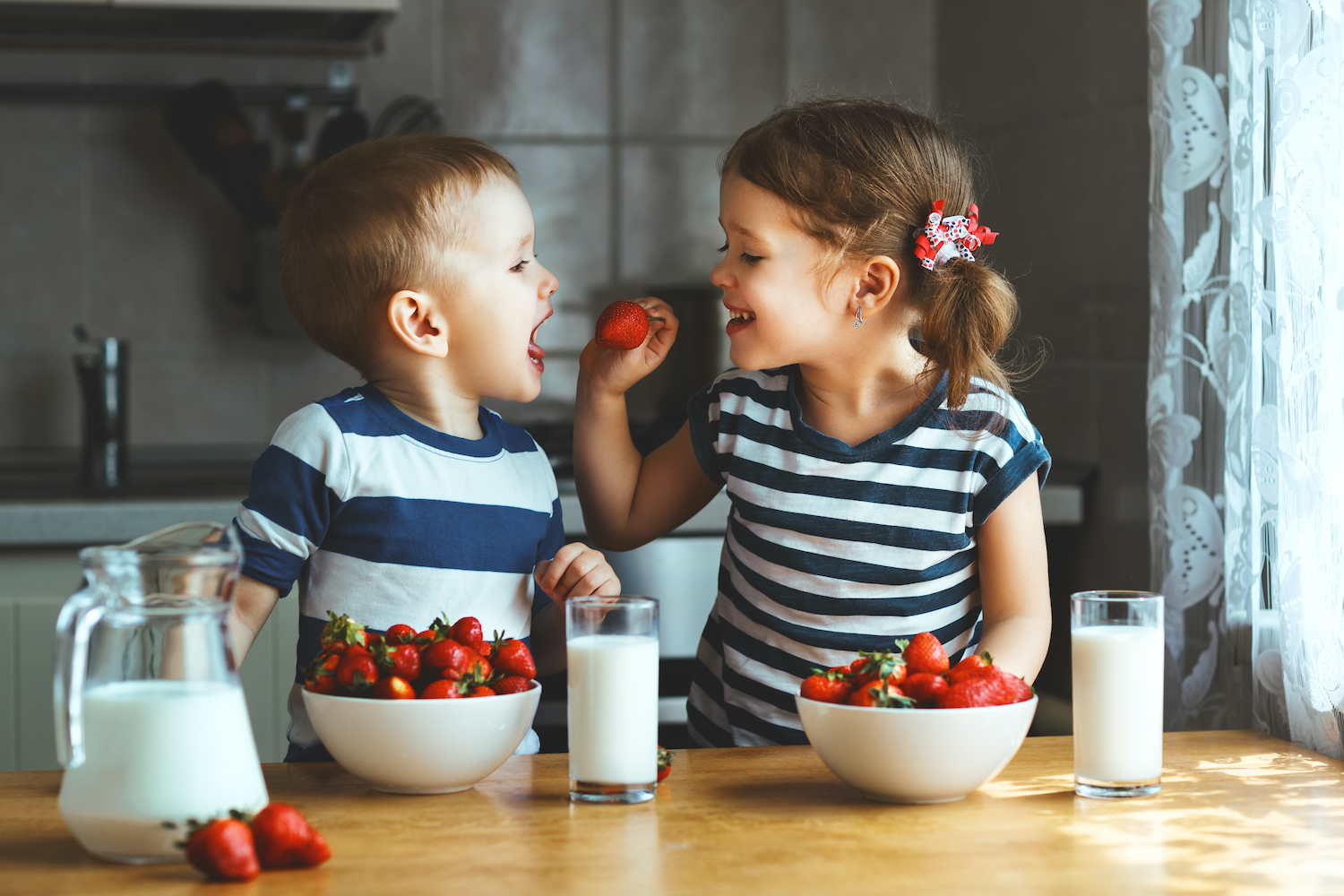 a brother and sister smile as they eat strawberries and milk in the kitchen