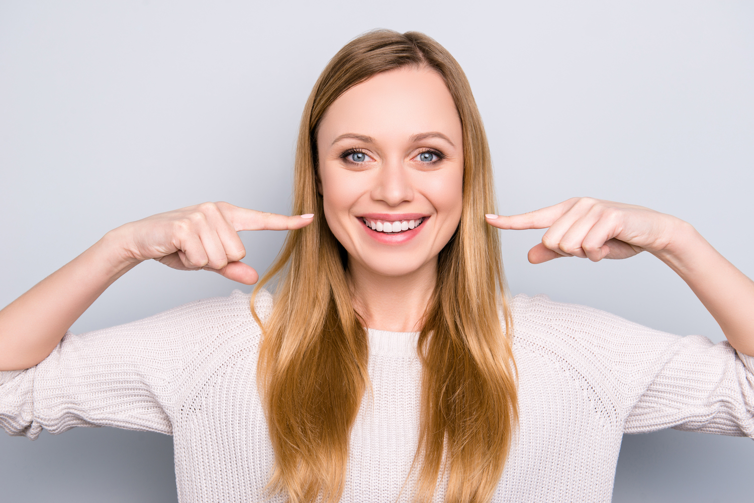 Blonde woman smiles while pointing to her white teeth
