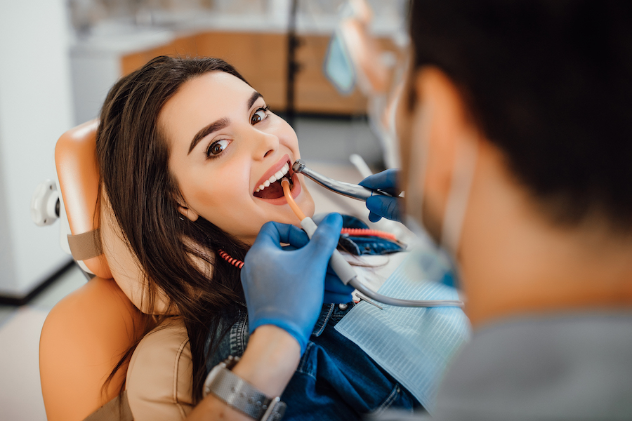 Young female patient visiting dentist office. Beautiful woman with healthy straight white teeth sitting at dental chair with open mouth during routine dental checkup while doctor working at teeth.